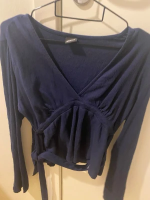 Mörkblå omlottopp från Gina Tricot - Snygg mörkblå omlottopp från Gina Tricot med långa ärmar och knyt i midjan. Toppen har v-ringad hals och är tillverkad i ett mjukt, strukturerat bomullsmaterial. Perfekt för dig som gillar stilrena och bekväma plagg med lite extra detalj.