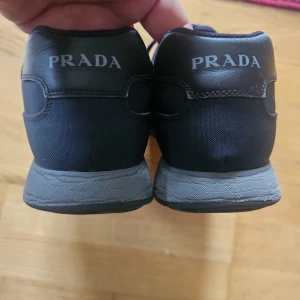 Svarta sneakers från Prada - Snygga svarta sneakers från Prada med klassisk logga på både plös och häl. Skorna har rund tå, platt grå sula och snörning framtill. Materialet är en mix av skinn och syntet, vilket ger en cool och stilren look. Perfekta för dig som gillar exklusiva märken och streetstyle.
