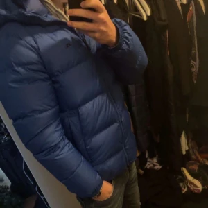 Blå pufferjacka från J Lindberg - Säljer en blå pufferjacka från J Lindberg med huva och dragkedja framtill. Jackan är vadderad och har en klassisk, sportig look som håller dig varm under kalla dagar. Perfekt för dig som gillar stilren stil nypris ca 5500kr. Fråga för mera bilder om behövs🙏