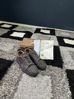 Christian Louboutin grå sneakers med nitar - Säljer ett par grå sneakers från Christian Louboutin i mocka med ikoniska röda sulor och coola nitar på tån. Skorna har snörning och en rund tå, vilket ger en edgy och lyxig vibe. Perfekta för dig som vill sticka ut med exklusiva detaljer.