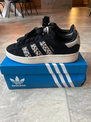 Adidas Campus 00s leopard sneakers - Svarta Adidas Campus 00s sneakers med beige detaljer och leopardmönstrade ränder på sidorna. Skorna har rund tå, platt sula och snörning framtill. Märkets logga syns på tungan och hälen. Ovandelen är i mocka och insidan är textil. Storlek 40 2/3 men passar mig som har storlek 40 vanligtvis.