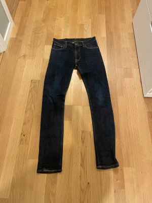 Mörkblå lean Dean Nudie Jeans - Snygga mörkblå jeans från Nudie Jeans med klassisk orange söm och logga på bakfickan. Modellen har smal passform, fem fickor och raka ben. Jeansen är tillverkade i slitstarkt denim och har en stilren look som funkar till sneakers eller boots.