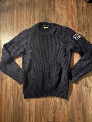 Mörkblå stickad tröja Jack & Jones - Mörkblå stickad tröja från Jack & Jones Premium med grov struktur och rund hals. Snygg detalj med patch på ena ärmen där det står 'Premium est. 1975'. Perfekt för kyliga dagar och enkel att matcha med jeans.