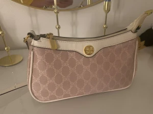 Beige axelväska från By Malene Birger - Snygg axelväska i beige med vitt mönster och vita detaljer från By Malene Birger. Väskan har guldfärgade metalldetaljer, justerbar axelrem och dragkedja upptill. Perfekt storlek för det viktigaste och ger en chic touch till din outfit.