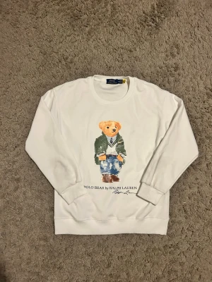 Vit sweatshirt Polo Bear Ralph Lauren - Vit sweatshirt från Polo Ralph Lauren med ikoniskt Polo Bear-tryck på framsidan. Tröjan har rund hals, långa ärmar och ribbade muddar. Materialet är mjuk bomull som känns skönt mot huden. Perfekt för dig som gillar klassisk och lekfull stil.