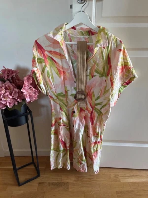 Blommig kortklänning från H&M - Fräsch kortklänning från H&M med tropiskt blommigt mönster i rosa, grönt och beige. Klänningen har krage, korta ärmar och knappar framtill. Medföljer ett beige bälte med rund spänne som markerar midjan. Perfekt för sommarens varma dagar. Använd en gång 