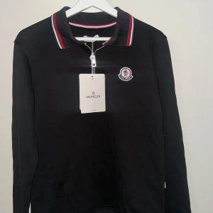 Svart pikétröja Moncler med half zip - Svart långärmad pikétröja från Moncler med half zip och klassisk krage. Tröjan har Moncler-logga på bröstet och snygga rödvita ränder på krage och muddar. Perfekt för dig som gillar stilrena och sportiga plagg med exklusiv känsla.