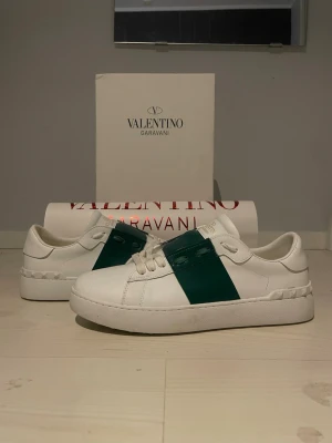 Valentino Opens - Snygga vita sneakers från Valentino Garavani med bred grön rem över ovansidan och ikoniska nitar på hälen. Skorna har snörning och platt sula, tillverkade i skinn för en lyxig känsla. Perfekt för dig som gillar stilrena och exklusiva sneakers. Box ingår 