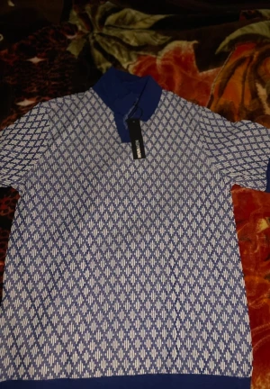 Blå och vit polotopp från New Moon - Säljer en snygg polotopp från New Moon i blått och vitt med ett rutigt mönster. Toppen har korta ärmar och en klassisk polokrage i marinblått. Perfekt för dig som gillar stilrena och grafiska detaljer.