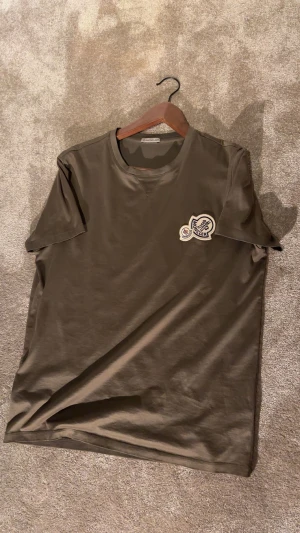 Olivgrön Moncler t-shirt med patch - Snygg olivgrön t-shirt från Moncler i mjuk bomull. Klassisk rund hals och korta ärmar. På bröstet sitter två broderade Moncler-patchar i vitt, blått och rött som ger en exklusiv vibe. Perfekt för dig som gillar stilrena designer med en twist.
