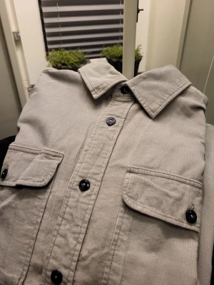Beige manchester overshirt Jack & Jones XL - Säljer en beige overshirt från Jack & Jones i manchester med svarta knappar och två stora bröstfickor. Insidan har ett rutigt foder i blått och vitt. Perfekt för lager på lager och ger en chill vibe till din outfit.