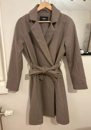 Beige kappa med bälte från Bik Bok - Snygg beige kappa från Bik Bok i klassisk modell med krage och knytbälte i midjan. Kappan har långa ärmar, knappar på insidan och en slits baktill för extra rörelsefrihet. Perfekt för dig som gillar stilrena och tidlösa plagg.