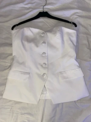 Vit axelbandslös korsettopp med knappar - Säljer en elegant vit korsettopp i satinliknande material. Toppen är axelbandslös och har klädda knappar framtill samt två dekorativa fickor. Insidan är fodrad för extra komfort och passformen är tight och smickrande. Perfekt för dig som vill sticka ut med en chic och clean look.