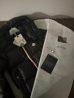 Svart Moncler pufferjacka med huva - Svart pufferjacka från Moncler med glansig finish, huva och klassisk logga på bröstet. Jackan har dragkedja med röd och vit rand längs öppningen, samt två sidofickor med dragkedja. Perfekt för kalla dagar och riktigt snygg streetstil.