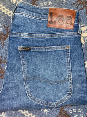 Blå skinny jeans från Lee - Snygga blå jeans från Lee med klassisk femficksdesign och brunt läderpatch med logga bak. Jeansen har en skinny passform och är tillverkade i mjukt denimtyg med lätt tvättad look. Perfekta för dig som gillar en smal siluett och stilren design.
