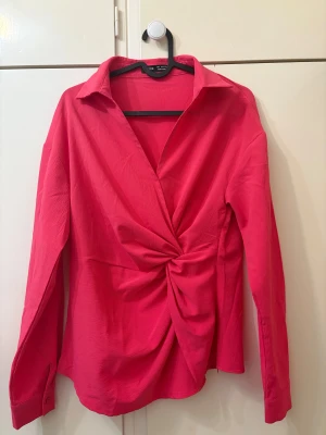 Rosa blus  - Det är en ceriserosa blus med långa ärmar och en skjortkrage. Framtill har den en knut som gör att tyget faller i fina veck. Ärmarna har enkla manschetter, och tyget är lite skrynkligt i strukturen. 