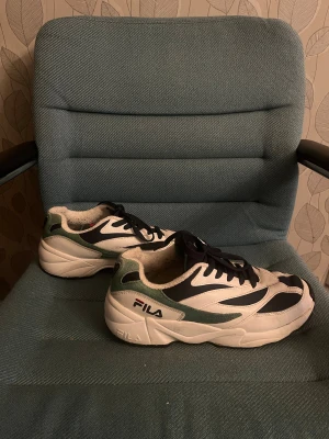 Fila sneakers i vitt, svart och grönt - Chunky Fila sneakers med cool retrostil i vitt, svart och grönt. Skorna har svart snörning, rund tå och en tjock vit sula. Ovandelen är i en mix av syntet och mocka, med Fila-logga på sidan och hälen. Perfekta för dig som gillar streetwear och sportig vibe.