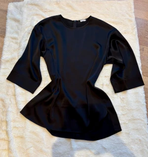 Svart blus Filippa K - Svart lite glansig stilren blus. Perfekt skick 10/10. Oranga kjolen som syns på bilderna är också till salu
