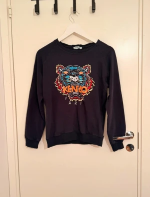 Svart Kenzo tröja med tigerbrodyr - Svart tröja från Kenzo med ikonisk tigerbrodyr i färgglada nyanser framtill. Klassisk rund halsringning och långa ärmar. Mjuk bomullskvalitet och ribbade muddar vid ärmslut och nederkant. Perfekt statement-piece för din garderob.