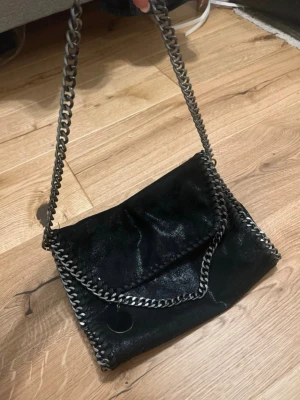 Svart axelväska med kedjedetaljer - Snygg svart axelväska med glansig finish och silverfärgade kedjor längs kanterna och som axelrem. Väskan har en mjuk, 💕