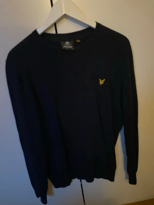 Marinblå stickad tröja Lyle & Scott - Snygg marinblå stickad tröja från Lyle & Scott med rund hals och gul broderad logga på bröstet. Tröjan har långärmad passform och är tillverkad i mjukt material som känns skönt mot huden. Perfekt för dig som gillar stilrena och klassiska plagg.