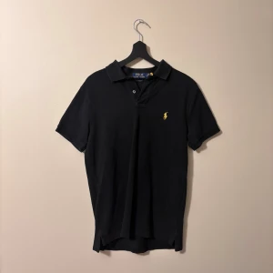 Svart piké (S) Polo Ralph Lauren - Svart pikétröja från Polo Ralph Lauren med klassisk krage och korta ärmar. Custom slim fit-modell i mjuk bomull. På bröstet finns den ikoniska gula broderade polospelaren. Perfekt för en clean och stilren look. Skickas säkert!