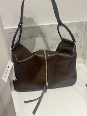 Wera Väska - Snygg mörkbrun axelväska från Wera med guldfärgad dragkedja och stilren design. Väskan har bred axelrem och medföljande innerficka i matchande färg. Perfekt för dig som gillar klassiska och tidlösa accessoarer.