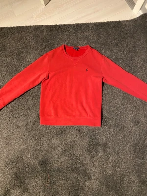 Röd crewneck från Polo Ralph Lauren - Röd crewneck-tröja från Polo Ralph Lauren med klassisk broderad logga på bröstet. Tröjan har rund halsringning, långa ärmar och är tillverkad i mjuk bomull. Perfekt för dig som gillar stilrena och tidlösa plagg. Storlek 14/16(L) år motsvarar ungefär S i herrstorlek.