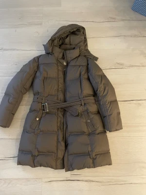 Ljusbrun lång dunjacka från Esprit - Säljer en brun lång dunjacka från Esprit med huva och bälte i midjan. Jackan har quiltad design, dragkedja och knappar framtill samt två stora fickor med knappstängning. Perfekt för kalla dagar och riktigt mysig att bära.