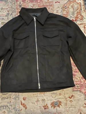 Svart overshirt med dragkedja XXL - Svart overshirt i storlek XXL med klassisk krage och två bröstfickor med lock. Jackan har en rak passform och stängs med en silverfärgad dragkedja framtill. 