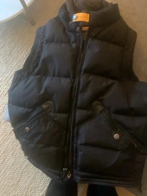 Svart dunväst från Parajumpers - Svart dunväst från Parajumpers Masterpiece Series med hög krage och orange detalj vid halsen. Västen har två stora fickor med dragkedja och tryckknappar, quiltad design och robust dragkedja framtill. Perfekt för kalla dagar och snygg att styla över hoodie.