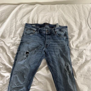 Blå slitna jeansbyxor - Säljer ett par blå jeansbyxor med slitningar och hål på benen. Klassisk femficksmodell med normal passform och knappgylf. Storlek W32 L30. Perfekta för en avslappnad och trendig look.