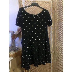 Svart prickig kortklänning - Supersöt svart klänning med vita prickar. Modellen är kort med rund halsringning och korta ärmar. Kjolen har en lätt utställd form som ger en skön och lekfull vibe. Perfekt för dig som gillar retrostil och vill sticka ut lite extra.