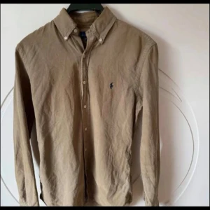 Beige skjorta från Ralph Lauren - Klassisk beige skjorta från Ralph Lauren med button-down krage och broderad logga på bröstet. Skjortan har långa ärmar och knäppning framtill. Perfekt för en stilren och avslappnad look. 