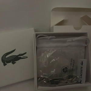 Silverfärgat armband från Lacoste - Säljer helt nytt och slutsålt online. Snyggt silverfärgat armband från Lacoste med en unik krokodildetalj. Perfekt för att ge en stilren touch till din outfit.