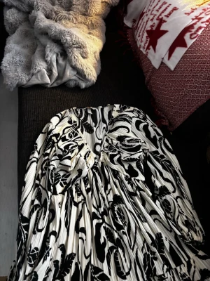 Vit och svart långklänning med v-ringning - Säljer en vit och svart långklänning med djup v-ringning och plisserad struktur. Klänningen har ett blommigt mönster och långa ärmar med lätt puff. Materialet känns mjukt och följsamt, perfekt för dig som gillar statement-plagg med coola detaljer.