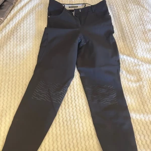 Svarta ridbyxor från Horka Equestrian Pro - Snygga svarta ridbyxor från Horka Equestrian Pro med diskreta mönster i form av prickar och linjer på benen. Byxorna har en praktisk ficka med dragkedja på sidan och dekorativa detaljer i vitt. Perfekta för dig som gillar ridning och vill ha stil på stallet.