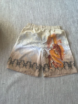 Beige shorts med drake och tiger - Säljer ett par beige badbyxor med elastisk midja och knäppning. Framsidan har en stor, färgstark print med drake och tiger i gult, orange och svart. Runt benen finns tribal-inspirerade svarta mönster. Materialet känns lätt och passar perfekt för varma dagar. Badbyxorna har aldrig blivit använda.