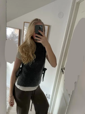 Grå stickad väst från & Other Stories - Mörkgrå stickad väst från & Other Stories med hög hals och knytband i sidorna. Västen är ärmlös och har en rak, loose passform. Perfekt att styla över en t-shirt eller skjorta för en trendig lager-på-lager-look. Prislapp finns kvar, skriv för fler bilder (: