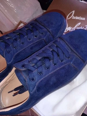 Blå mocka sneakers från Christian Louboutin - Säljer ett par snygga sneakers från Christian Louboutin i marinblå mocka med rund tå och platt sula. Skorna har klassisk snörning och stilren design, perfekt för dig som gillar exklusiva märken och vill sticka ut med färg. Kommer med originalkartong och dustbag.