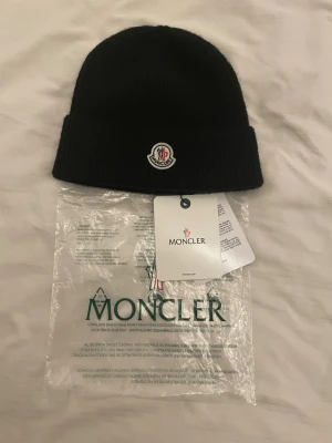 Svart Moncler mössa i ull - Säljer en svart Moncler mössa i mjuk och ribbstickad ull med klassisk uppvikt kant och broderad logotyp framtill. Perfekt för dig som vill ha en clean och stilren look under kyliga dagar. Mössan kommer med originalförpackning och tags.