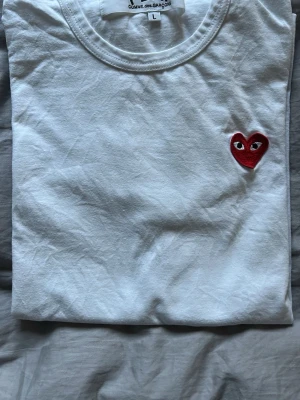 Vit t-shirt från Comme des Garçons PLAY - Säljer en clean vit t-shirt från Comme des Garçons PLAY i storlek L. T-shirten har ett broderat rött hjärta med ögon på bröstet och klassisk rund hals. Materialet är mjuk bomull och modellen är kortärmad, perfekt för dig som gillar stilrena plagg med en twist. Nypris 949kr
