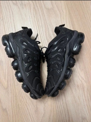Nike Air Vapormax Plus svarta sneakers - Nike Air Vapormax Plus sneakers i helsvart färg med chunky, genomskinlig sula och snygga vågformade detaljer på ovansidan. Skorna har snörning och en platt, rund tå. Ovandelen är i syntetmaterial och mesh för extra komfort. Perfekt för dig som gillar sportig och modern stil.