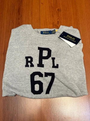 Grå tröja från Polo Ralph Lauren - Oandvänd snygg grå tröja från Polo Ralph Lauren med svart tryck 'RPL 67' på bröstet. Tröjan har rund hals, ribbade muddar och är långärmad. Materialet är mjuk bomull som känns skönt mot huden. Perfekt för dig som gillar stilrena plagg. Storlek xs. Passar 170 - 180cm. Original pris 2400kr. 