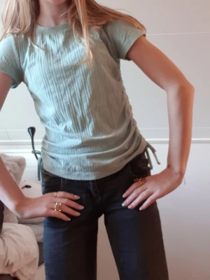 Ljusgrön ribbad topp med knyt - Säljer en ljusgrön ribbad topp med korta ärmar och snygga knytband i sidorna. Toppen har en rund halsringning och är i ett mjukt, stretchigt material som sitter bekvämt. Perfekt till jeans eller kjol för en fräsch look.