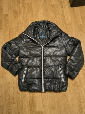 Svart pufferjacka från On The Peak - Säljer en svart pufferjacka från On The Peak med glansig finish och vita dragkedjor. Jackan har hög krage, två fickor med dragkedja och quiltad design. Perfekt för kalla dagar och riktigt skön att bära.