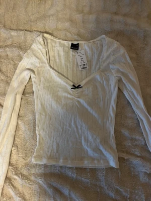 Vit topp med rosett Gina Tricot - Supersöt vit långärmad topp från Gina Tricot i pointelle-mönster. Toppen har v-ringning och en liten svart rosett framtill. Tillverkad i mjuk bomullsmix och har smala ribbade ränder samt spetskant vid halsen.