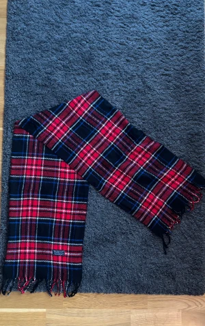 Burberry Halsduk (ÄKTA) - Snygg halsduk från Burberry i klassiskt rutig design med röda, svarta, blå och vita toner. Mjuk och varm, perfekt att vira runt halsen. Fransar i ändarna ger en sotifistikerad vibe.  ✅ Skick: 8,5/10 🧵Material: Fin ull 🎨 Färg: flerfärgad, primärt röd (svart, blå vit) ✨Väldigt skön, tidlös och snygg scarf med fint och välbehandlat material   ‼️Skickas samma dag eller dagen efter köpet genomförs‼️