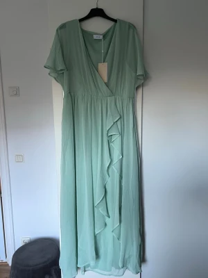 Ljusgrön långklänning från Vila - Superfin ljusgrön långklänning från Vila med omlott framtill och volangdetalj. Klänningen har korta ärmar och ett luftigt, flowy tyg.