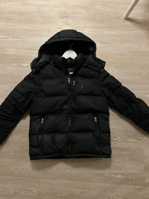 Svart pufferjacka från Polo Ralph Lauren - Svart pufferjacka från Polo Ralph Lauren med huva, två sidofickor med dragkedja och en röd logga på bröstet. Jackan har quiltad design och är perfekt för kalla dagar. Stängs med dragkedja framtill och har en modern, normal passform.